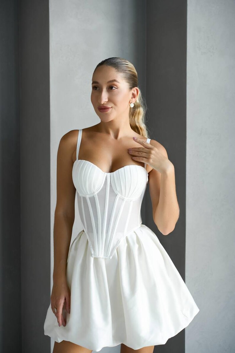 May include: A white corset-style top with a structured bodice and thin straps, paired with a matching mini skirt. The dress features a fitted silhouette and a flared skirt, creating a cohesive and elegant look. The outfit is suitable for special occasions.