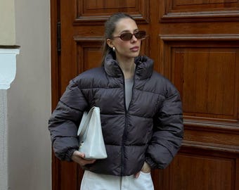 May include: A dark brown puffer jacket with a high collar and a zipper closure. The jacket is paired with white pants and a white handbag. The person is wearing sunglasses and earrings. The background is a wooden door.