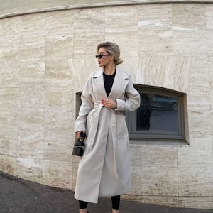 Peut inclure: Un long manteau beige clair avec une ceinture assortie, porté sur un haut et un legging noirs. Le manteau a un design classique avec de larges revers et des poches. La tenue est complétée par des talons hauts noirs et un petit sac à main noir.