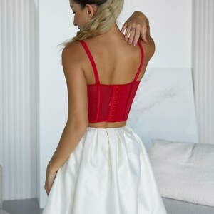 May include: A red corset top with thin straps and a hook-and-eye closure, paired with a white satin mini skirt. The outfit is a stylish ensemble, perfect for a special occasion. The skirt has a voluminous, bubble-like shape.