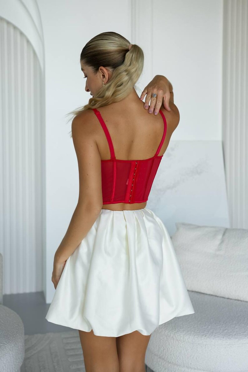 May include: A red corset top with thin straps and a hook-and-eye closure, paired with a white satin mini skirt. The outfit is a stylish ensemble, perfect for a special occasion. The skirt has a voluminous, bubble-like shape.