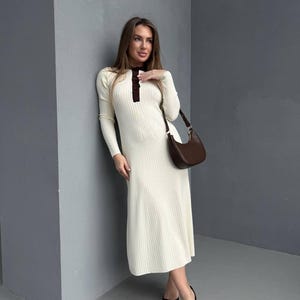 May include: Cream-colored, long-sleeve midi dress with a ribbed texture. The dress features a dark brown collar and button detail. A brown shoulder bag and black pointed-toe heels complete the outfit.