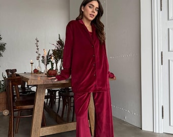 May include: A burgundy velvet pajama set, including a button-down shirt and wide-leg pants. The outfit is paired with black fuzzy slippers. The setting is a room with a wooden table and decorative greenery.