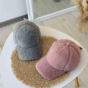 Peut inclure: Deux casquettes de baseball duveteuses, une grise et une rose, faites d'une matière douce et texturée. Elles sont présentées sur une table blanche, posées sur un tapis tissé doré.