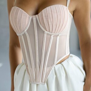 May include: A blush-colored corset with a structured bodice and delicate straps. The corset features vertical boning and a sheer mesh panel, paired with a white skirt. The design is elegant and form-fitting, suitable for special occasions.