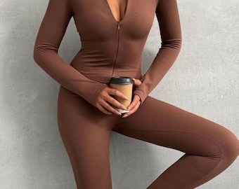 May include: A brown ribbed jumpsuit with a zip-up front and long sleeves. The model is wearing white sneakers and holding a coffee cup. The jumpsuit is form-fitting and appears to be made of a stretchy material.