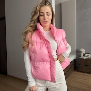 Puede incluir: Un chaleco acolchado rosa brillante con cuello alto. El chaleco se usa sobre una camisa blanca de manga larga y pantalones blancos. El chaleco tiene una textura brillante y suave y un diseño acolchado.