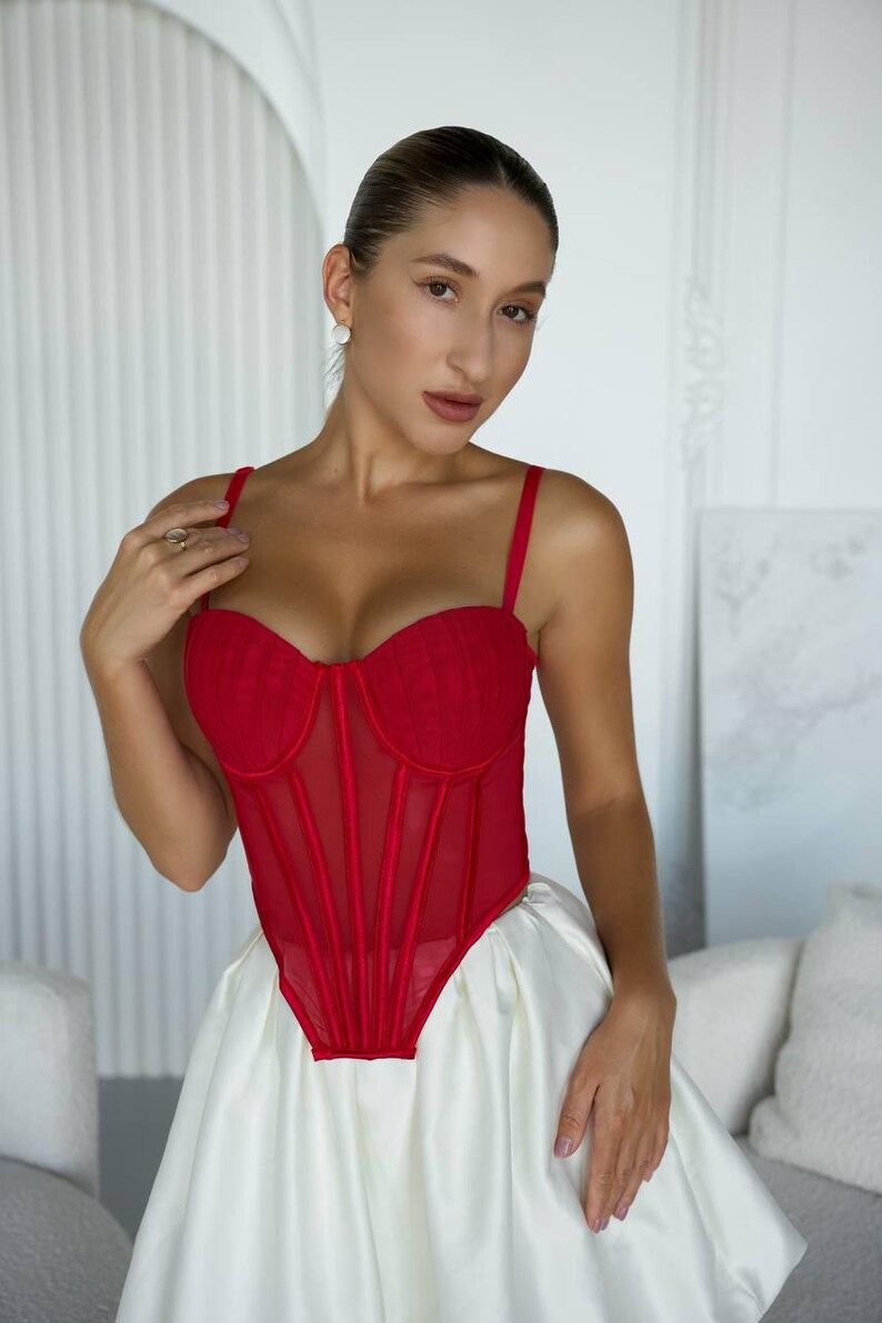 May include: A red corset top with spaghetti straps and a sheer mesh panel. The bodice has a structured design with vertical seams. The top is paired with a white satin skirt, creating a contrasting color palette.