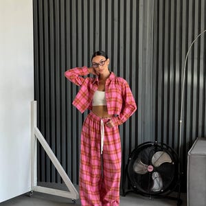 May include: A pink and brown plaid matching set, consisting of a cropped button-down shirt and wide-leg pants. The pants have a drawstring waist. The outfit is paired with a white crop top and white sneakers. The background features a dark gray vertical striped wall.