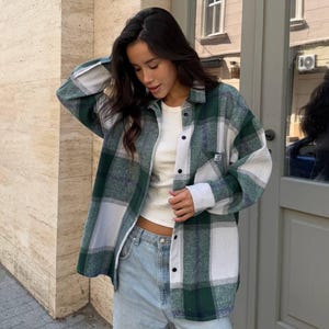 May include: A green, white, and gray plaid flannel shirt worn over a white top and light blue jeans. The shirt has a button-down front and a small pocket. The outfit is casual and comfortable.