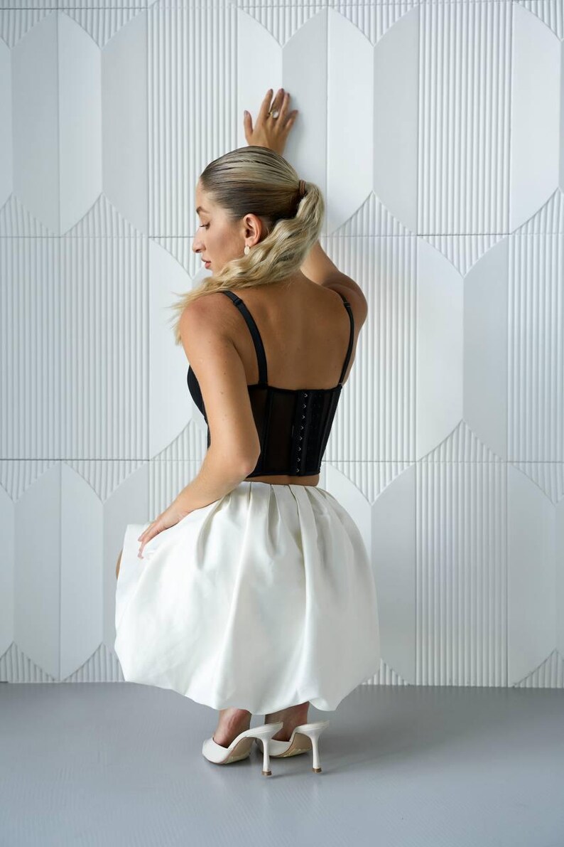 May include: A black corset top with adjustable straps and a white, knee-length skirt. The outfit is paired with white high-heeled shoes. The background features a white geometric wall.