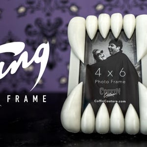 May include: A white fang-shaped photo frame with a 4x6 photo. The frame has a black and white photo of a couple. The text "Fang FRAME" is in white, and the website "CoffinCouture.com" is visible.