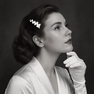 May include: A black and white image of a woman with dark hair styled in waves, wearing a white satin robe and white gloves. A white, wavy hair clip adorns her hair. The woman is looking up, with her chin resting on her gloved hand.