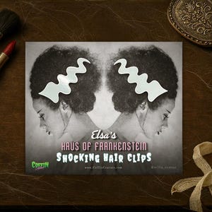 May include: An advertisement for Elsa's Haus of Frankenstein Shocking Hair Clips. The image features a black and white graphic of a woman with a white zig-zag hair clip, makeup brushes, a lipstick, and a vintage hand mirror.