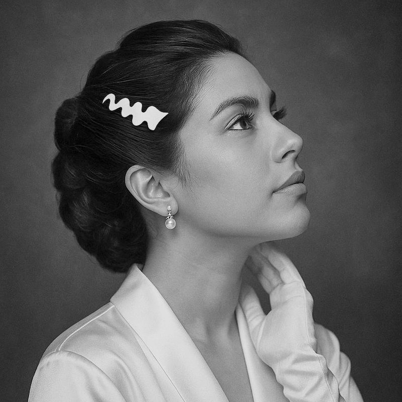 May include: Black and white image of a woman with dark hair styled in an updo. She wears a white hair clip with a wavy design, a pearl earring, and a white satin robe. The woman is looking upwards.