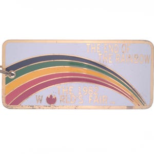 May include: A gold-colored metal keychain with a white background and a rainbow arch. The text "The End of the Rainbow" is at the top and "The 1982 World's Fair" is at the bottom. A red flame-like symbol is next to the word "World's".