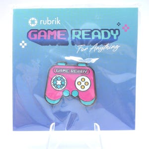 May include: A pink and teal video game controller pin with the words "GAME READY" on it. The pin is on a blue background with the words "rubrik" and "GAME READY For Anything".