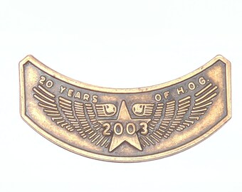 Peut inclure: Un emblème incurvé de couleur bronze avec le texte "20 YEARS OF H.O.G." et l'année "2003". L'emblème présente une étoile centrale et des ailes stylisées.