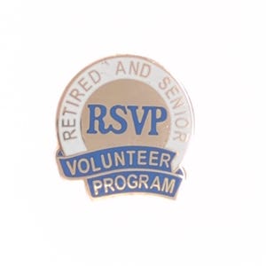 Peut inclure: Épinglette dorée avec bordure blanche et accents bleus. Le texte "RETIRED AND SENIOR" est inscrit en haut, et "RSVP" en bleu au centre. En dessous, une bannière bleue indique "VOLUNTEER PROGRAM".