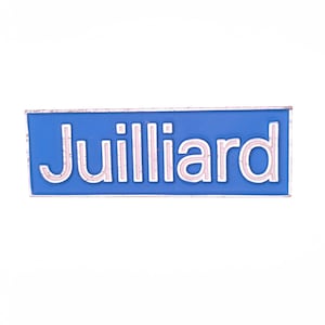 May include: A rectangular blue enamel pin with the word "Juilliard" in white capital letters. The pin has a white border and is set against a white background.