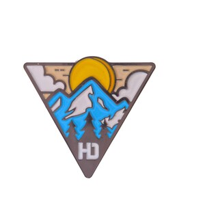 May include: A triangular enamel pin featuring a mountain landscape with a yellow sun, blue mountains, and green trees. The pin has a brown border and the letters "HD" at the bottom.