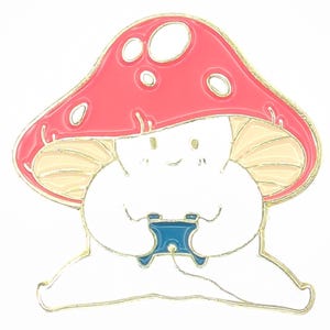 May include: A mushroom-shaped enamel pin with a red cap and white spots. The pin has a white body and a blue controller. The pin has a gold outline and a smiling face. This cute pin is perfect for adding a touch of fun to any outfit.