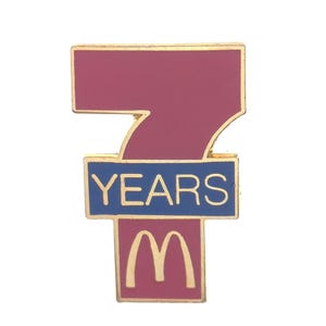 May include: A gold-colored pin with a burgundy number seven, a blue banner with the text "YEARS" and a burgundy McDonald's logo.