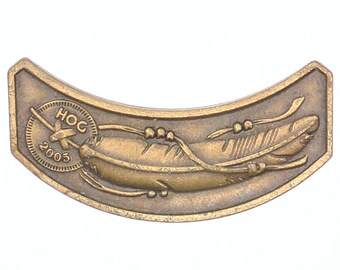 Peut inclure: Un emblème métallique incurvé de couleur bronze, représentant une plume et le texte "HOG 2003". L'emblème présente un design détaillé avec un élément circulaire et une plume stylisée, probablement un objet commémoratif.