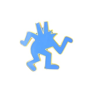 May include: A bright blue enamel pin featuring a stylized, abstract figure in a dynamic running pose. The figure has a jagged, multi-pointed head and is outlined in a thin, polished gold-tone metal border, creating a sharp contrast against the blue.