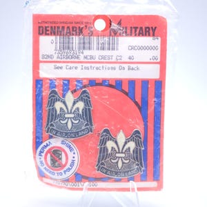 May include: Two military insignia pins on a red card. The pins feature a fleur-de-lis design with wings, the text "IN AIR, ON LAND", and the text "82ND AIRBORNE NCBU CREST C2". The card reads "DENMARK'S MILITARY".