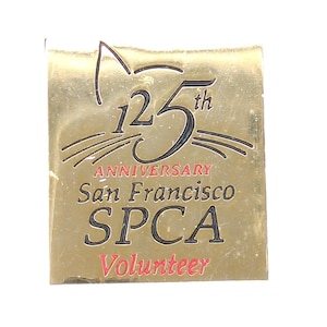 May include: A gold-colored rectangular pin with a cat silhouette at the top. The pin commemorates the 125th anniversary of the San Francisco SPCA. The word "Volunteer" is in red at the bottom.