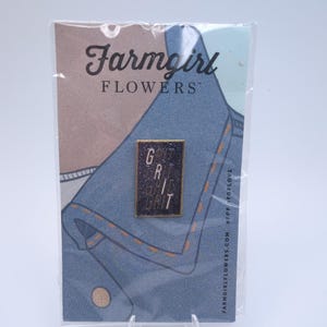May include: A pin with the words "GRIT" in a rectangular frame. The pin is on a card with the text "Farmgirl FLOWERS" and a denim-like design. The background is a light blue and tan color.