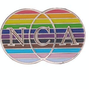May include: A silver-colored pin featuring two overlapping circles with a rainbow-striped design. The letters "NCA" are in the center of the pin. The pin has a shiny, metallic finish and a colorful, eye-catching design.