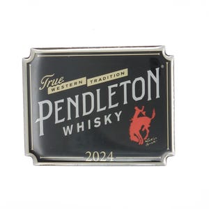 Peut inclure: Une boucle de ceinture rectangulaire en métal avec une finition émaillée noire et une bordure argentée. Le texte indique True Western Tradition Pendleton Whisky 2024. Un graphique rouge d'un cheval au galop est présent avec le texte Let'er Buck.