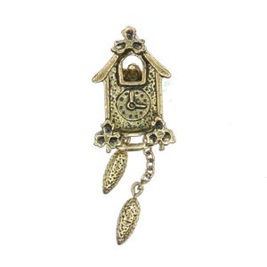May include: A gold-coloured cuckoo clock pendant with intricate detailing. The clock has a small bird, a clock face, and decorative features. Two cone-shaped weights are suspended from a chain beneath the clock.