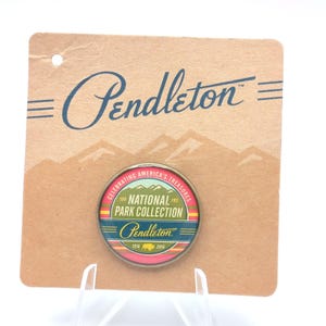 May include: A round National Park Collection pin with the Pendleton logo, celebrating America's treasures. The pin is on a cardboard backing with the Pendleton logo and mountain graphic. The pin features a colorful design.