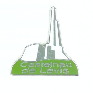 May include: A silver-colored enamel pin featuring a stylized depiction of a building or structure. The pin has a green base with the text "Castelnau de Lévis" in white. The design is simple and geometric.