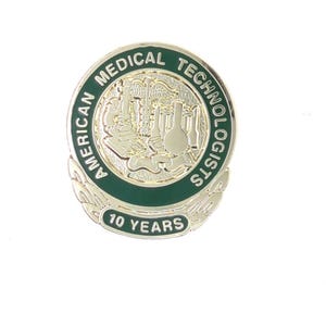 May include: A circular pin with a silver finish and a green border. The pin displays the text "AMERICAN MEDICAL TECHNOLOGISTS". The centre has a gold-coloured illustration of lab equipment. The bottom of the pin reads "10 YEARS".