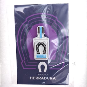 May include: A metal enamel pin shaped like a Tequila Herradura bottle, featuring a white bottle with a black horseshoe logo and blue accents. The pin is mounted on a dark purple backing card with a white horseshoe icon and the text Tequila Herradura.