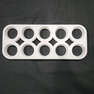 May include: A white plastic egg holder with ten circular compartments. The holder has a rectangular shape with rounded edges and diamond-shaped cutouts between the egg spaces. The background is a dark, textured surface.
