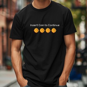 May include: Black t-shirt with the text "Insert Coin to Continue" in white and a row of four pixelated gold coins.