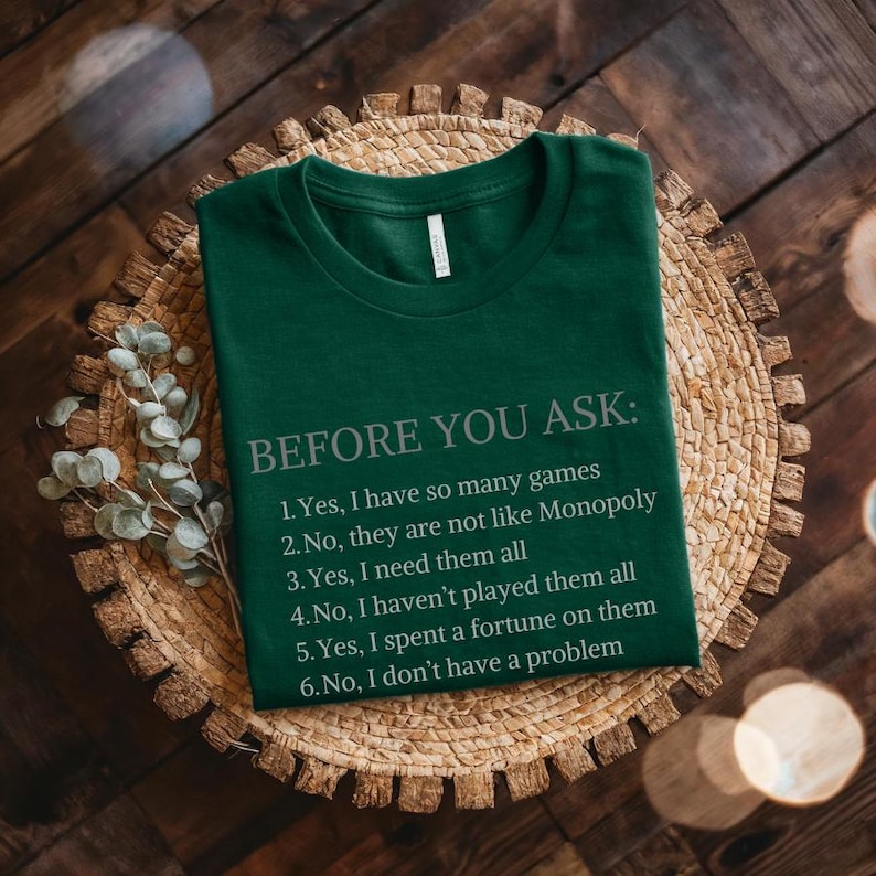 May include: A green t-shirt with the text "BEFORE YOU ASK:" and a list of six questions and answers about board games. The text is in a light gray color.