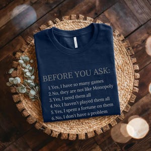 May include: A navy blue t-shirt with white text that reads "BEFORE YOU ASK:" followed by a list of six statements about board games. The statements are: "1. Yes, I have so many games", "2. No, they are not like Monopoly", "3. Yes, I need them all", "4. No, I haven't played them all", "5. Yes, I spent a fortune on them", and "6. No, I don't have a problem."