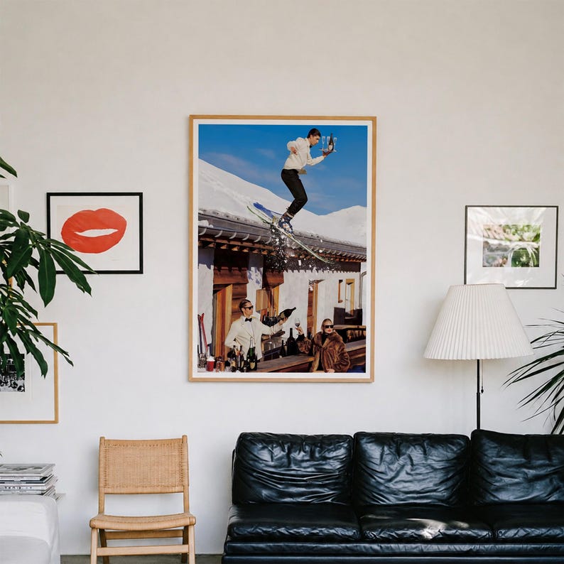 May include: A large framed wall art print featuring a surreal scene of a skier jumping over a rustic wooden building while holding a tray of champagne glasses, with two people in formal attire standing below on a terrace against a snowy mountain.