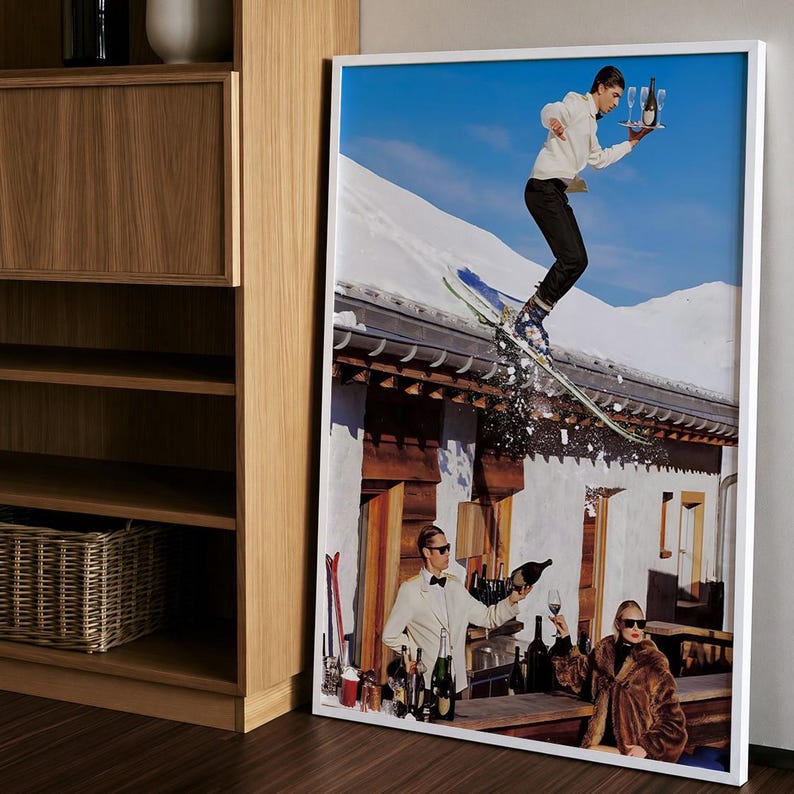 May include: A framed wall art print featuring a surreal scene with a skier jumping over a building roof while holding a tray of champagne, and below, a person in a white jacket pouring champagne for another person wearing a brown fur coat.