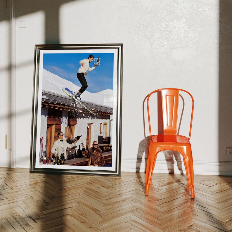 May include: A framed photographic print featuring a skier jumping over a building roof while holding a tray with champagne, with two people sitting at a bar below. Beside the frame stands a bright orange metal chair with a curved backrest.