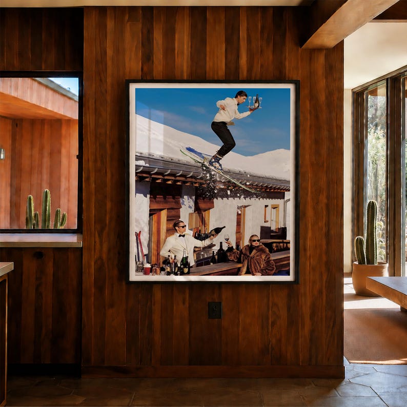 May include: A framed wall art print depicting a surreal scene with a person skiing off a snowy roof while holding a tray of champagne glasses, and two people in formal attire at a bar below, set against a bright blue sky and mountain backdrop.