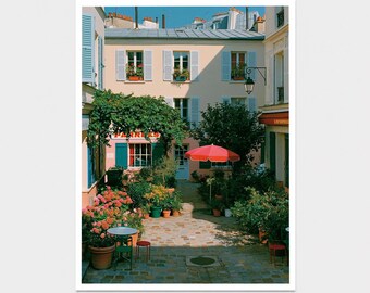 May include: A photographic print of a European courtyard featuring a cream-colored building with blue shutters, a bright red umbrella, and a cobblestone ground. Potted plants and flowers fill the space, and the text PARRES is visible.