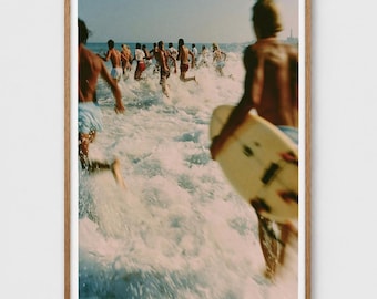 Può includere: Una stampa fotografica incorniciata che mostra un gruppo di persone che corrono tra le onde dell'oceano. In primo piano, una persona trasporta una tavola da surf chiara. La scena cattura onde bianche, acqua blu e un cielo limpido e soleggiato.