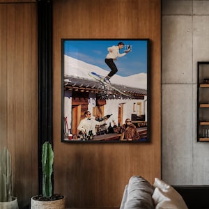 May include: A framed wall art print depicting a person in a white sweater and black pants skiing off a snowy roof while holding a tray with glasses, above two people at a bar counter, one in a white suit and one in a brown fur coat.